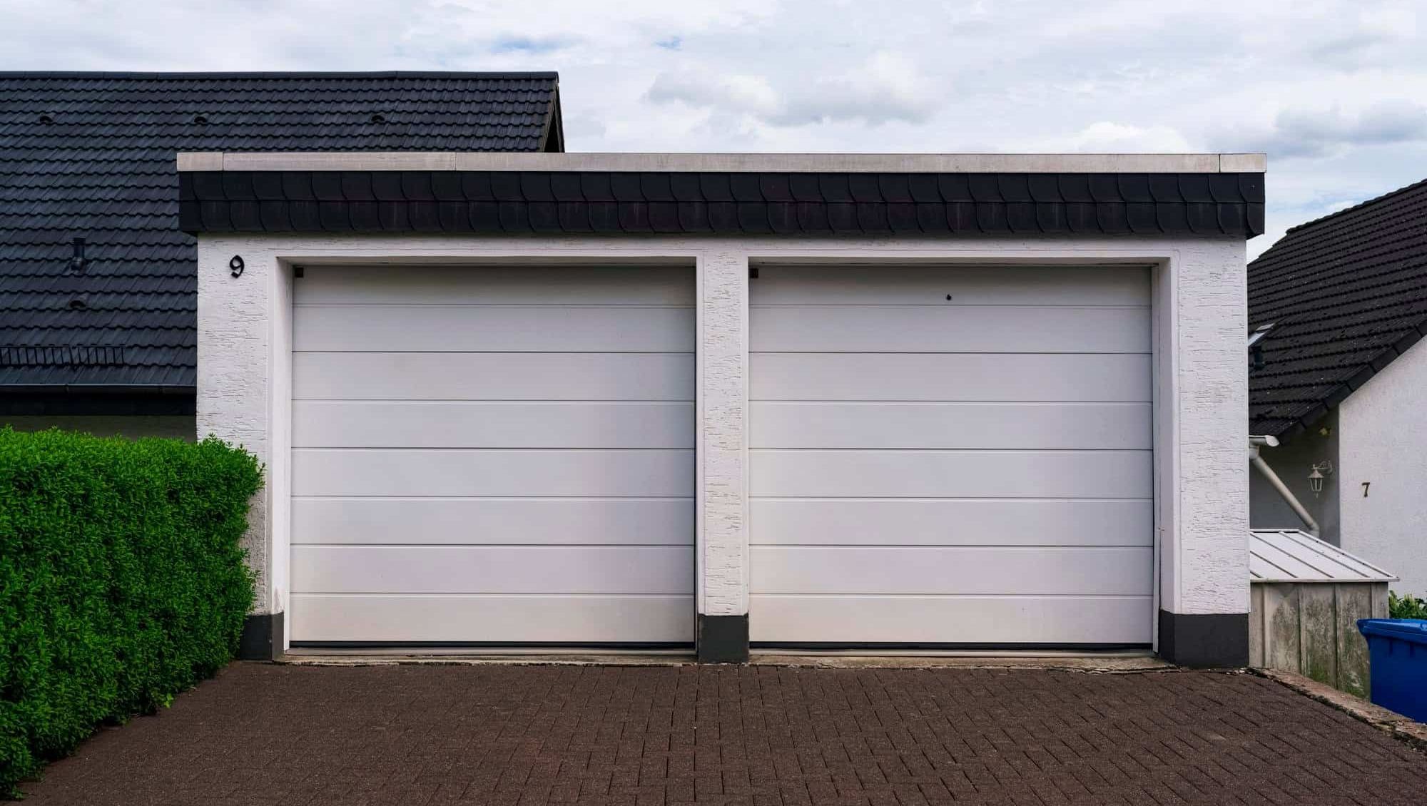 En dobbel garasje med hvite porter og mørkt tak, plassert ved innkjørsel til boligområde.