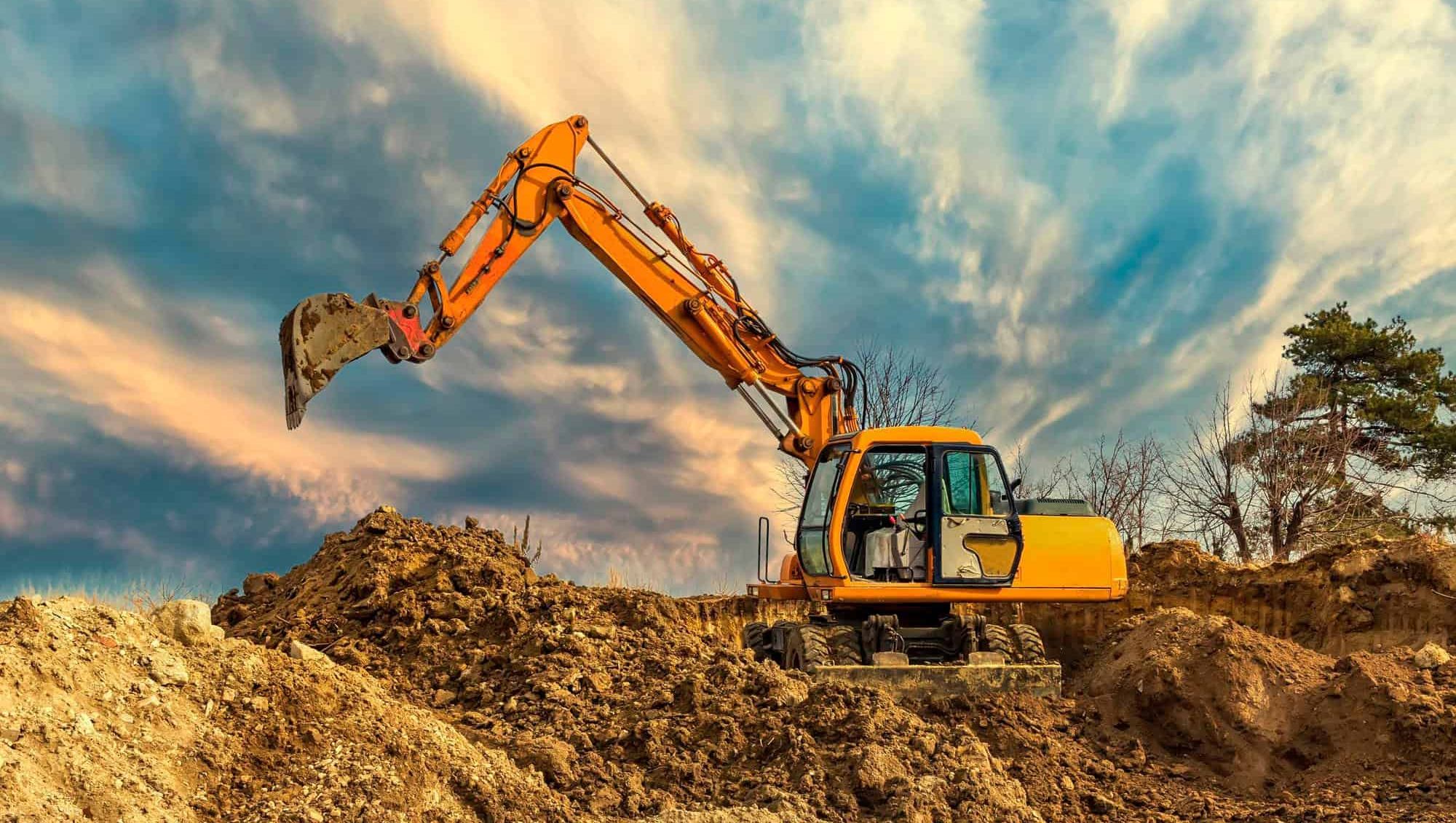 En gul gravemaskin arbeider på en byggeplass, omgitt av jordhauger under en dramatisk himmel.