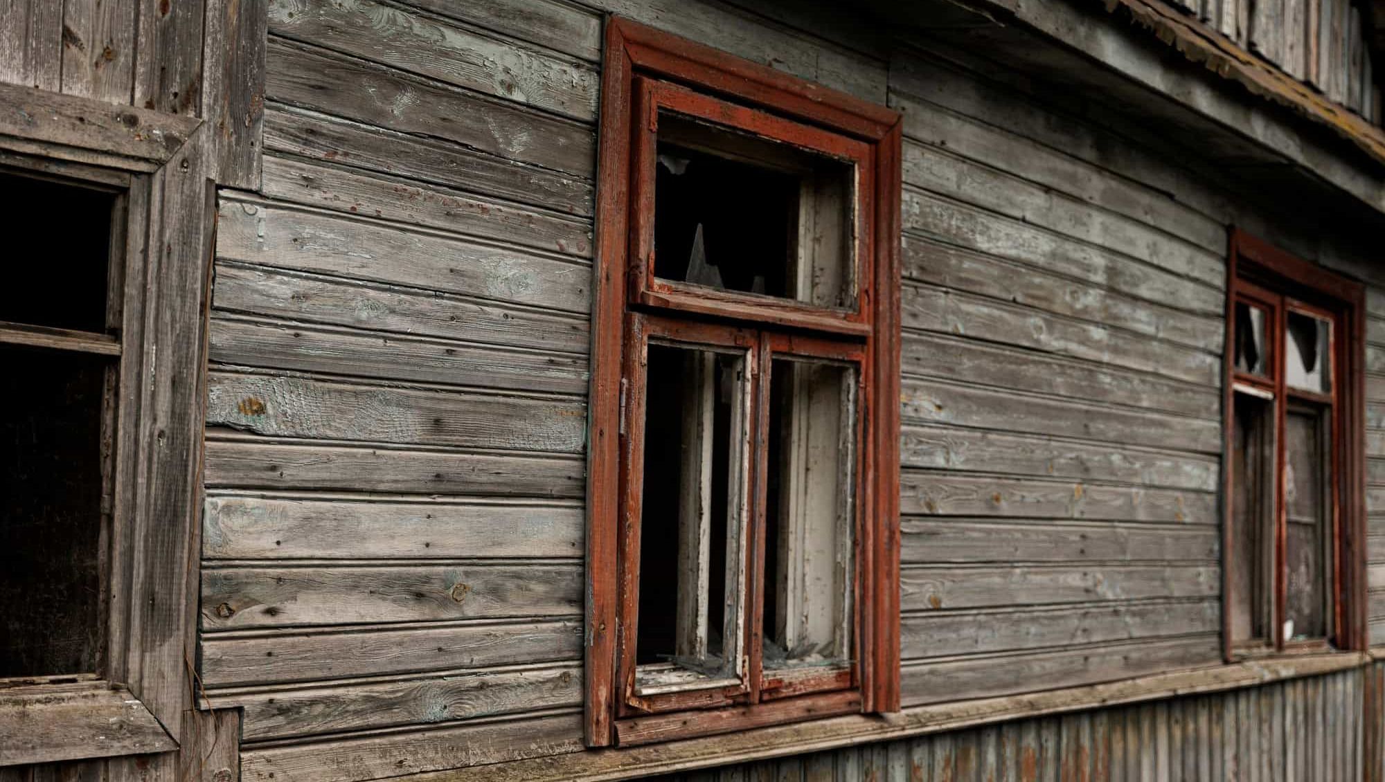 Fasade på et gammelt, slitt trehus med falmet panel og knuste vindusruter i røde rammer.