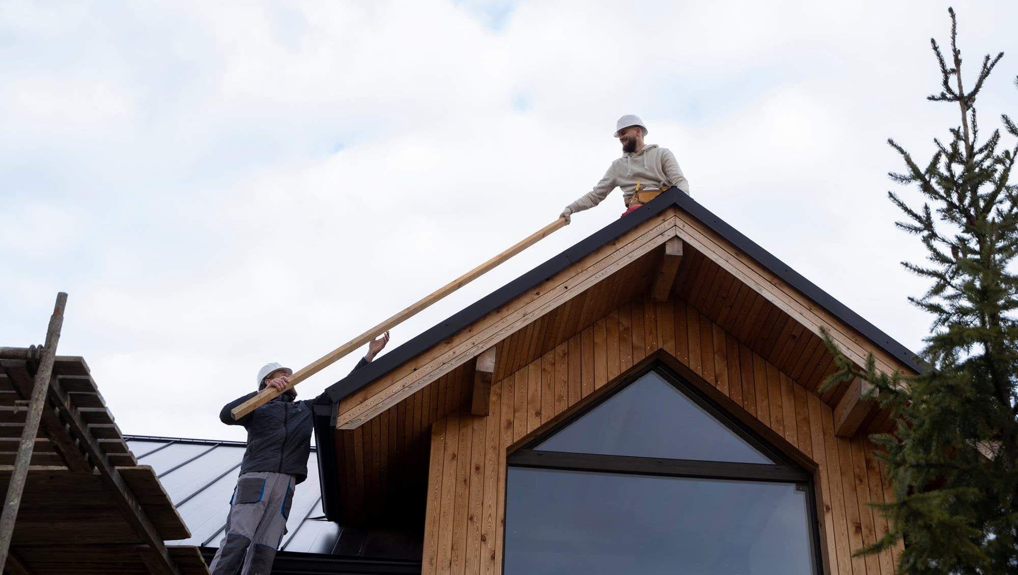 To bygningsarbeidere på taket løfter og overrekker en lang planke under husbygging.